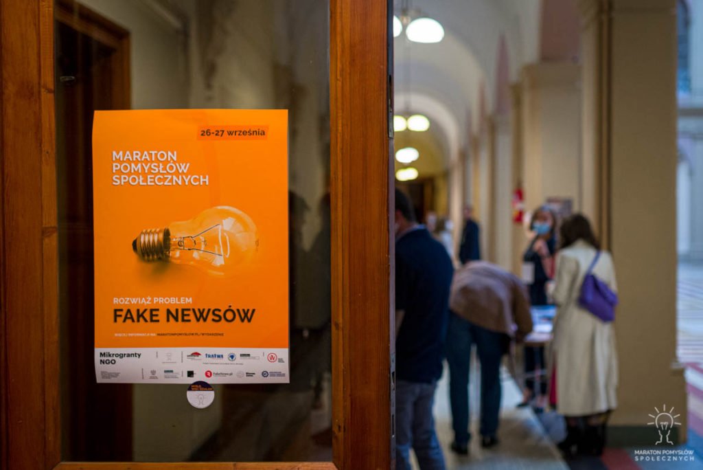 Plakat wydarzenia - Maraton Pomysłów Społecznych: Fake newsy na drzwiach. W tle goście wydarzenia w trakcie gali finałowej w NOT Wrocław.