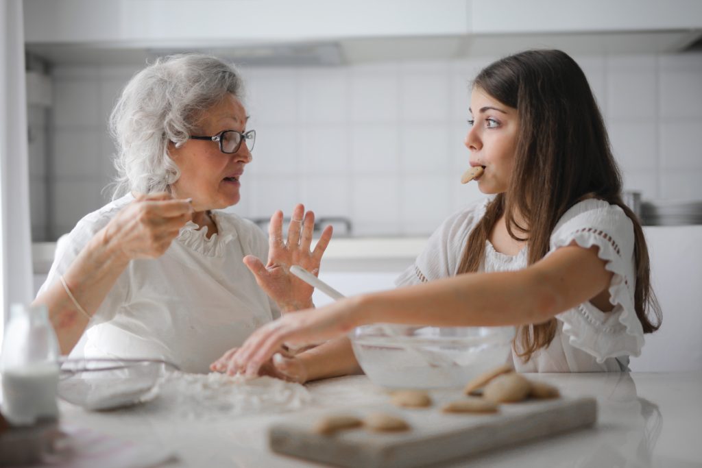 Babcia i wnuczka siedzące przy stole w trakcie gotowania. Wnuczka trzyma w ustach ciastko. Babcia tłumaczy jej coś.