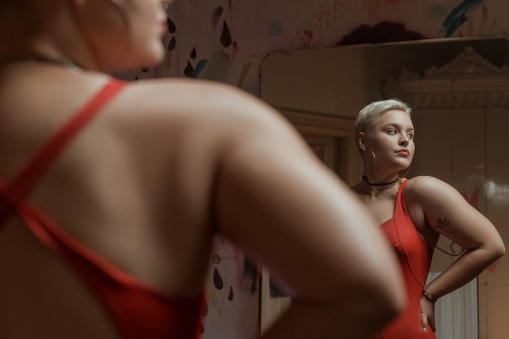 Kobieta w czerwonym kostiumie kąpielowym stojąca przed lustrem. Po prawej stronie w odbiciu widoczna jej twarz, po lewej zarys sylwetki.