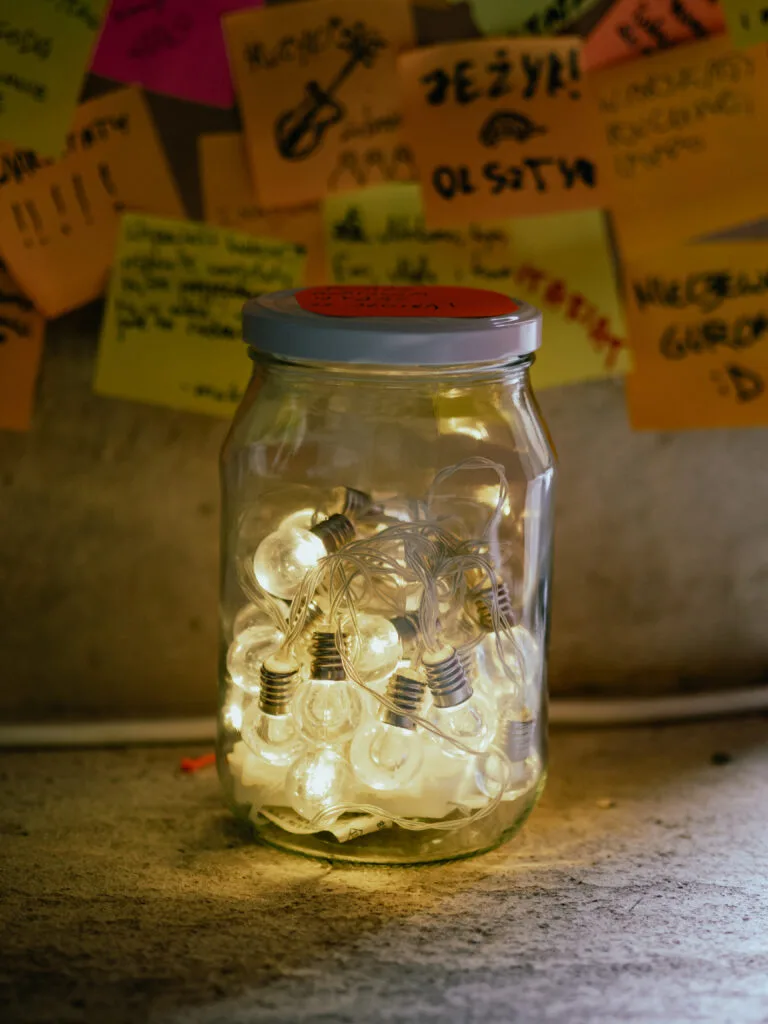 Słoik z łańcuchem świecących żarówek w środku. z tyłu widoczny fragment tablicy z ręcznie zapisanymi kolorowymi karteczkami.