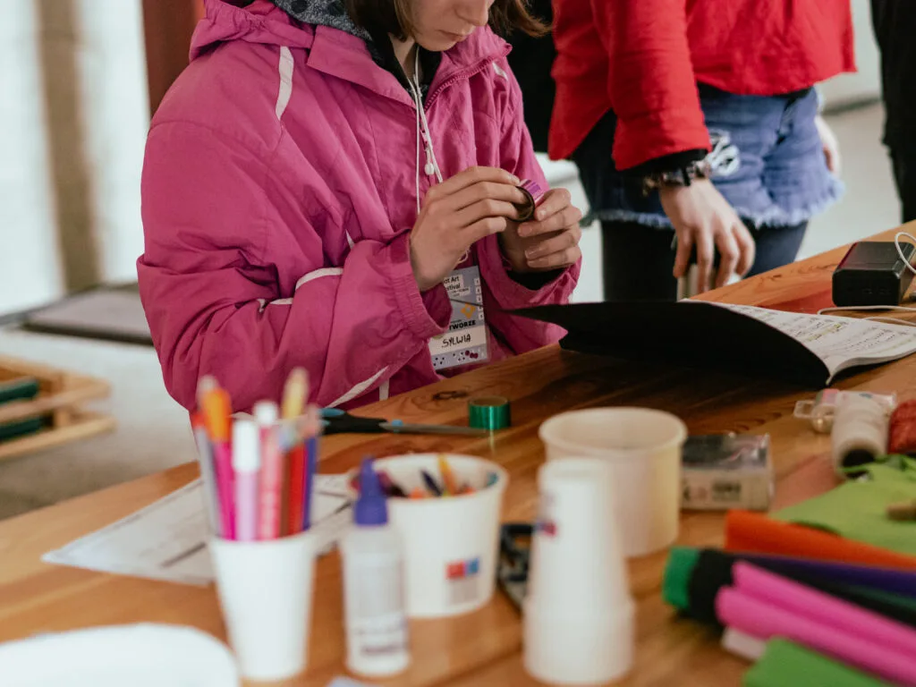 Dziewczyna w różowej kurtce w skupieniu odkleja taśmę przy stole z materiałami kreatywnymi.