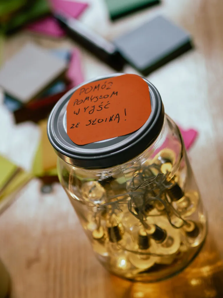 Szklany słoik z łańcuchem żarówek w środku. Na zakrętce napis na pomarańczowej kartce: pomóż pomysłom wyjść ze słoika.